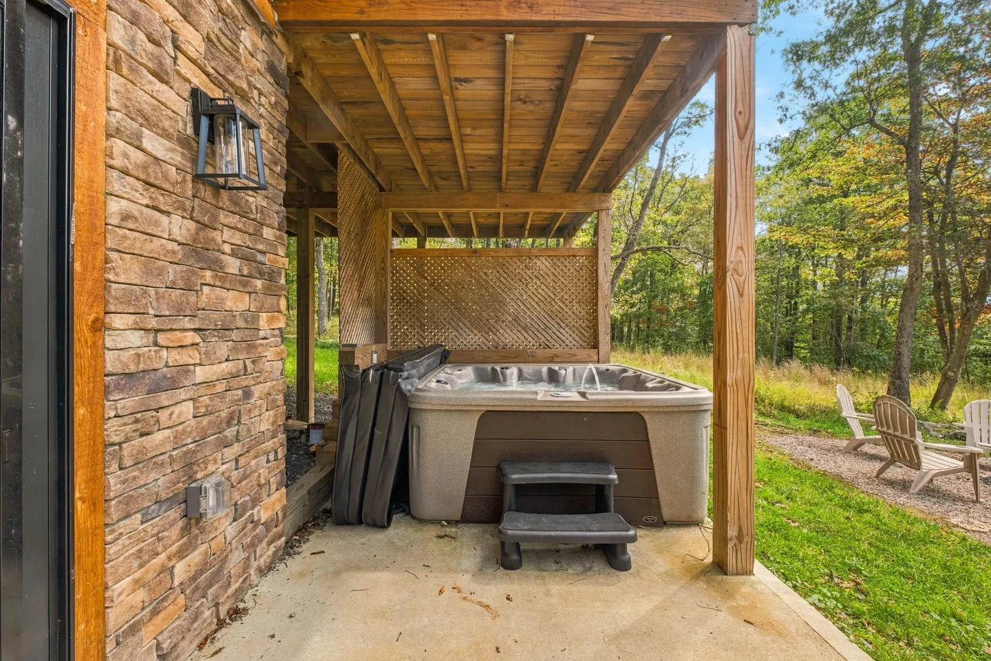 A hot tub under a covered patio next to a stone exterior wall with views of the lawn and trees.
