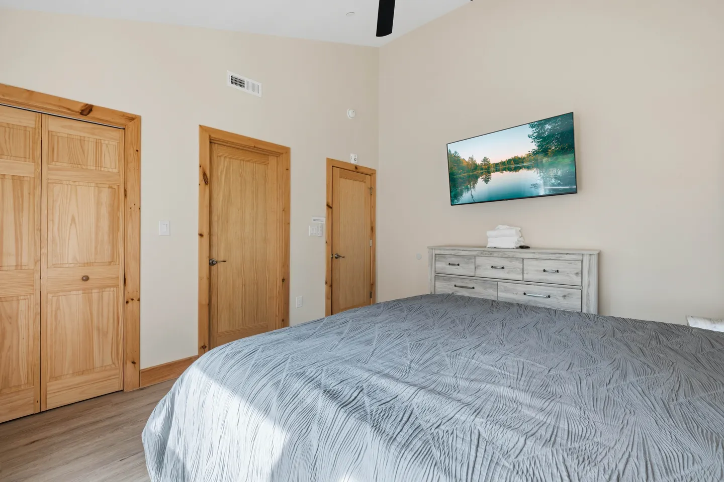 Bedroom featuring a made bed with a textured gray comforter, light wood doors, a white chest of drawers, and a wall-mounted flat-screen TV.