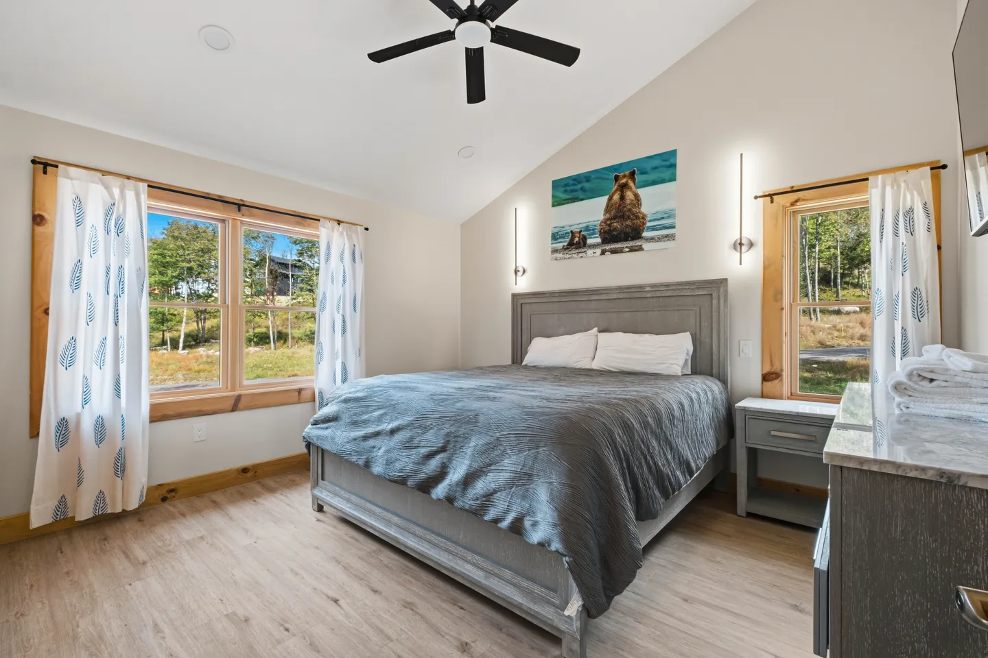 Bedroom with a wooden bed frame, grey bedding, vaulted ceiling with a fan, and two windows with patterned curtains.