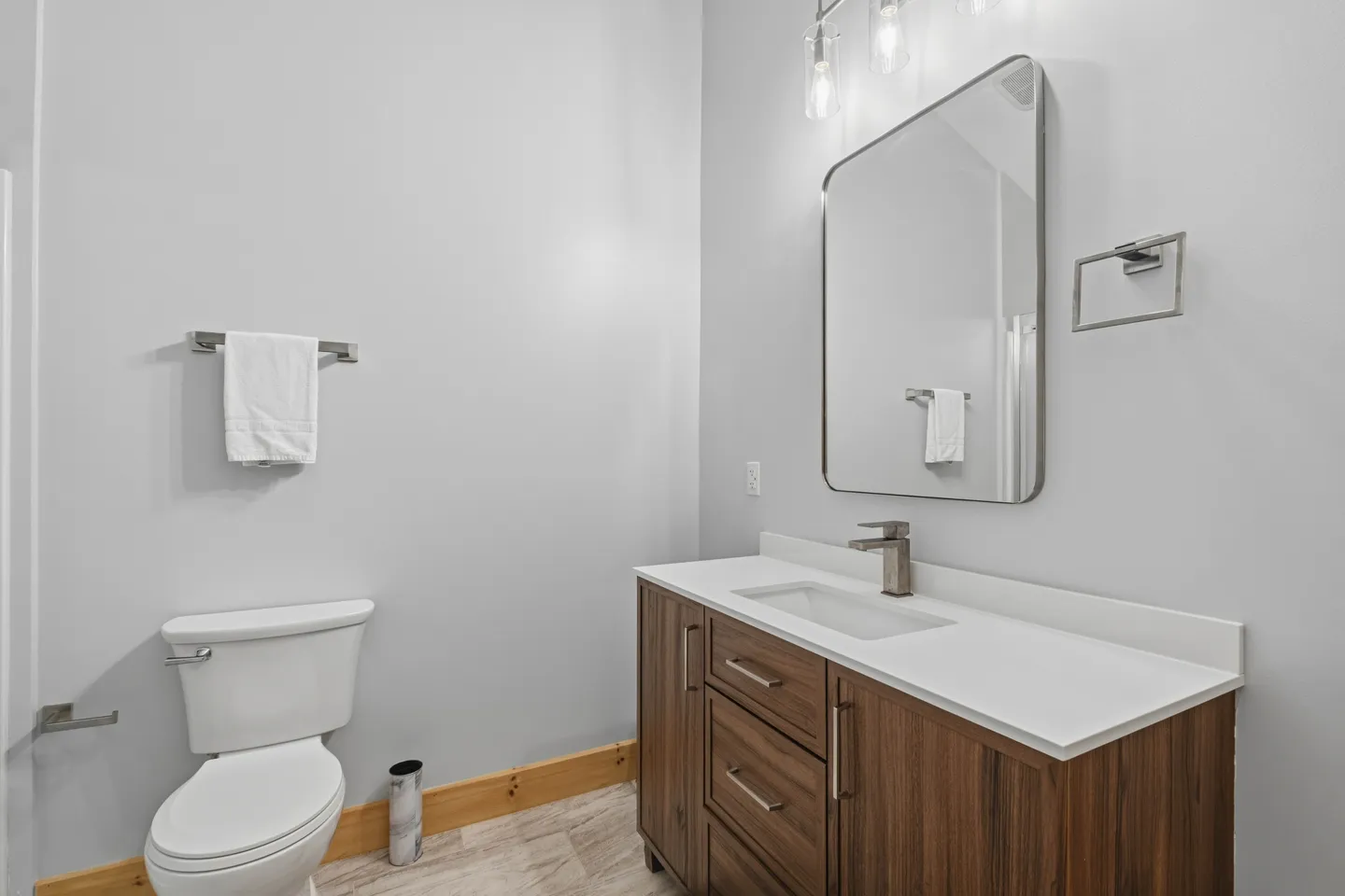 Bathroom with a wood-finish vanity, white countertop, modern mirror, and toilet against light gray walls.
