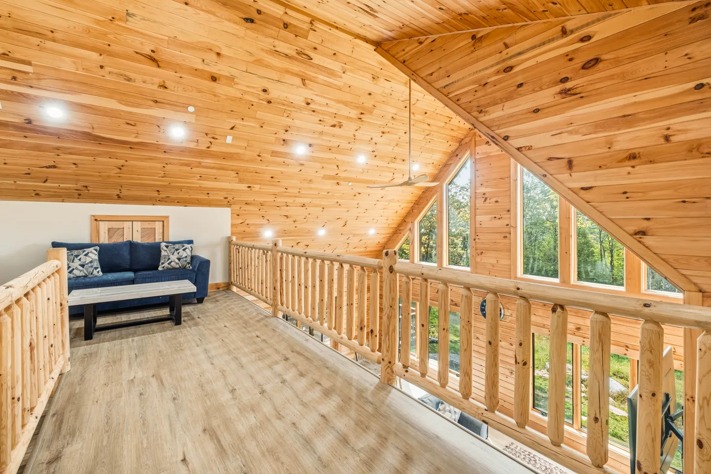 Upstairs loft area featuring wood paneled vaulted ceilings, log railings, light wood flooring, and a blue sofa.