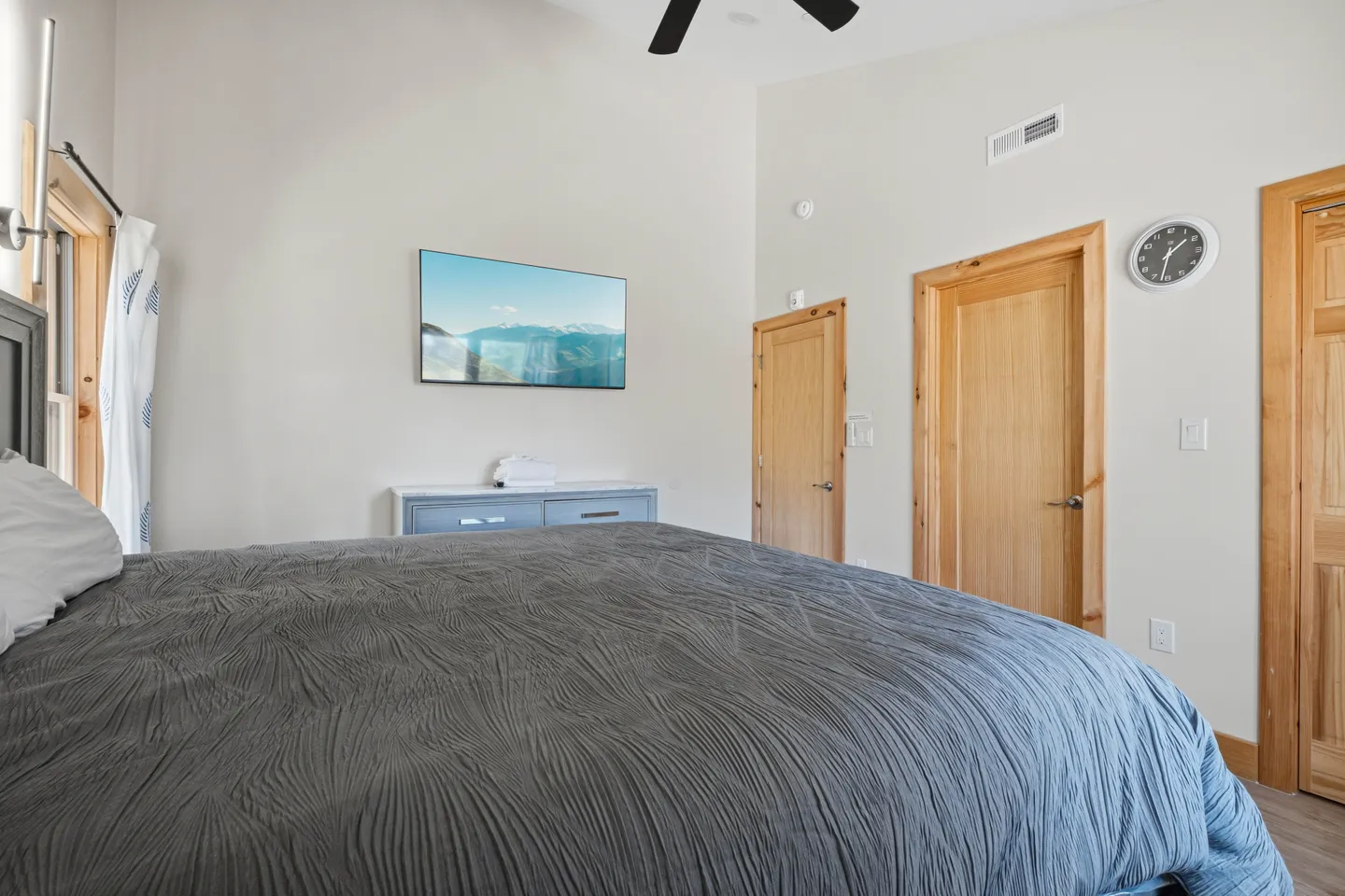Bedroom featuring a bed with a textured gray comforter, a wall-mounted TV displaying a mountain landscape, and light wood interior doors.