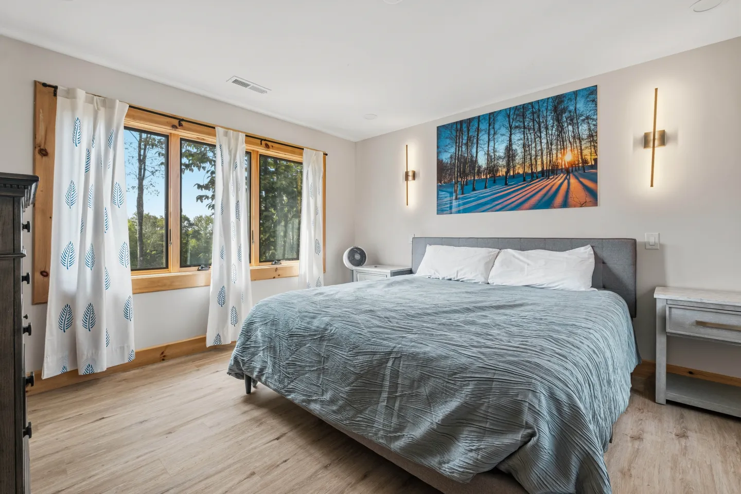 Bedroom with a large bed, nightstands, modern wall sconces, a winter landscape print, and large windows framed in wood.