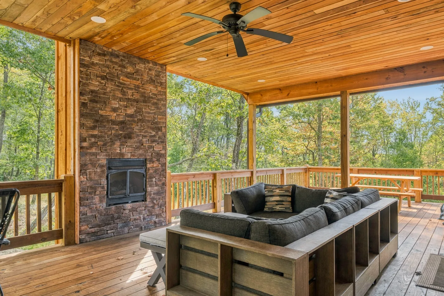 A covered deck with a stone fireplace, wood ceiling with a fan, outdoor sectional sofa, and railings overlooking surrounding green trees.