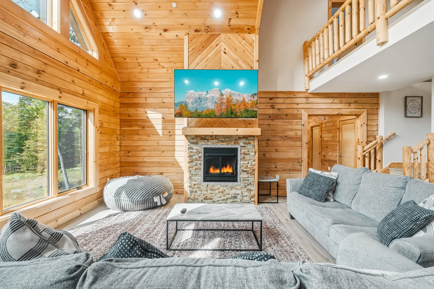 Living room with wood-paneled walls and ceiling, a stone fireplace, mounted TV, sectional sofa, and loft railing above.