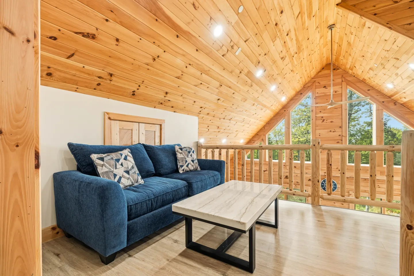 Upstairs loft sitting area with a blue couch, coffee table, and wood-paneled vaulted ceiling overlooking a lower level.