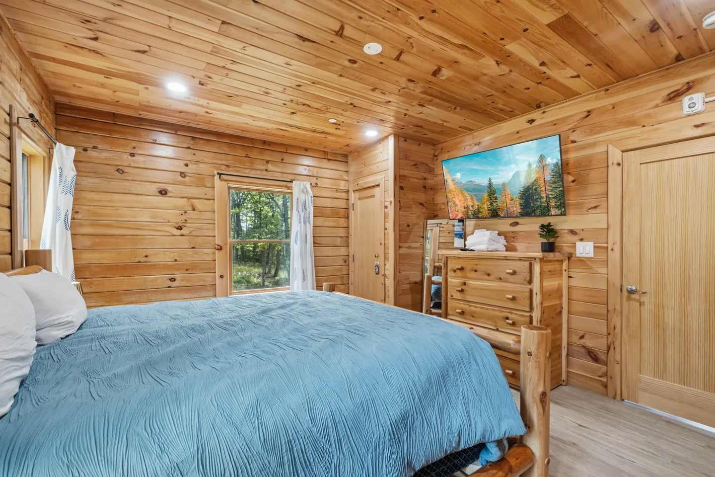 Bedroom with knotty pine wood-paneled walls and ceiling, log-frame bed with blue bedding, and a wall-mounted TV.