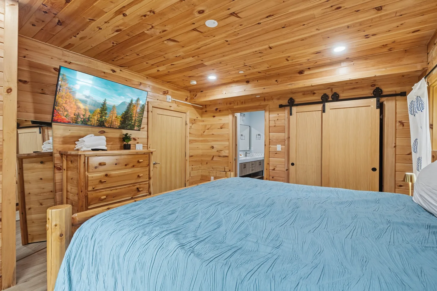 Bedroom with knotty pine wood-paneled walls and ceiling, featuring a bed with a textured blue coverlet, a wall-mounted TV, and a wooden dresser.