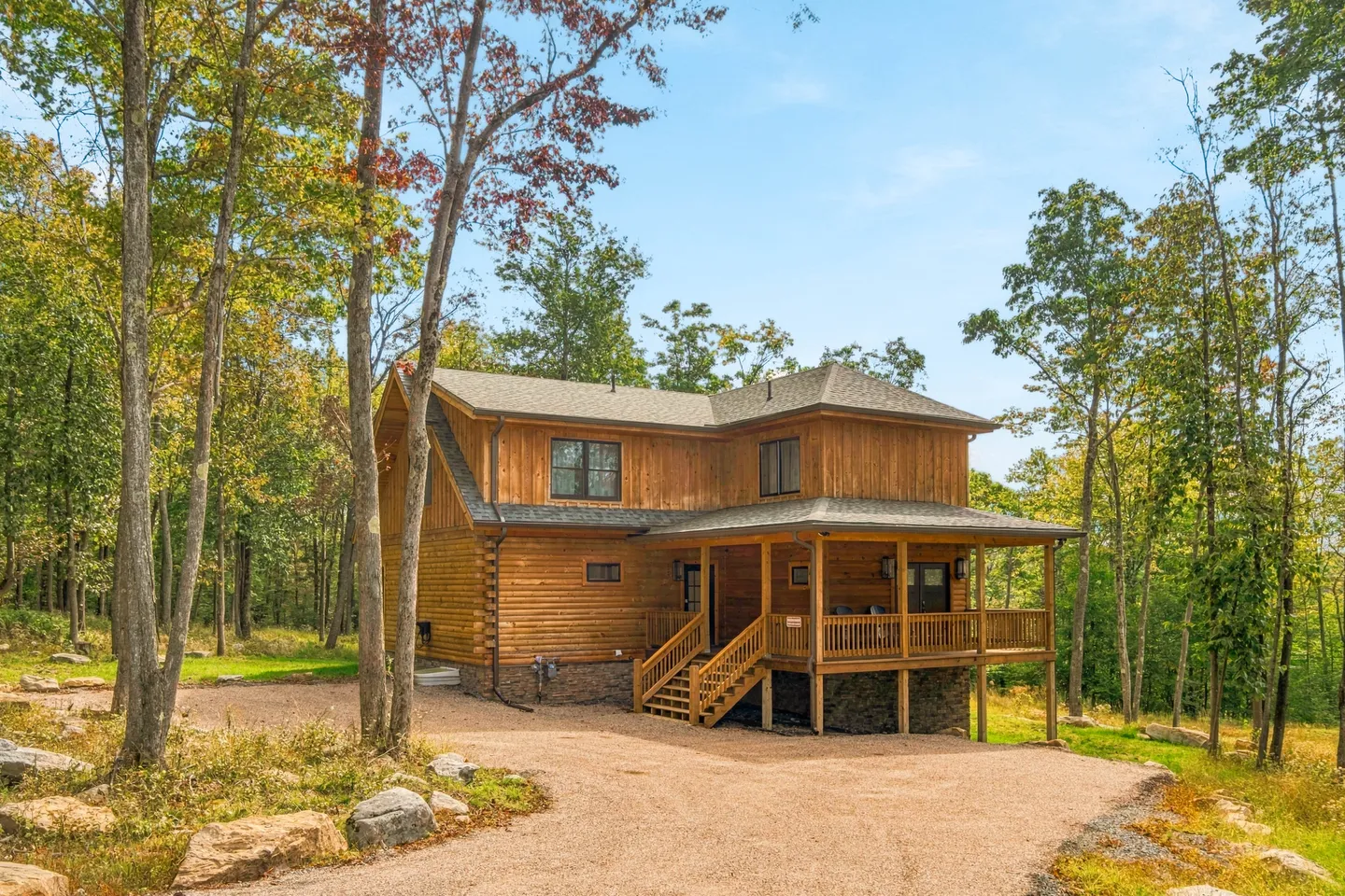 Exterior of a two-story log-style cabin with a covered wooden porch, set among trees along a gravel driveway.