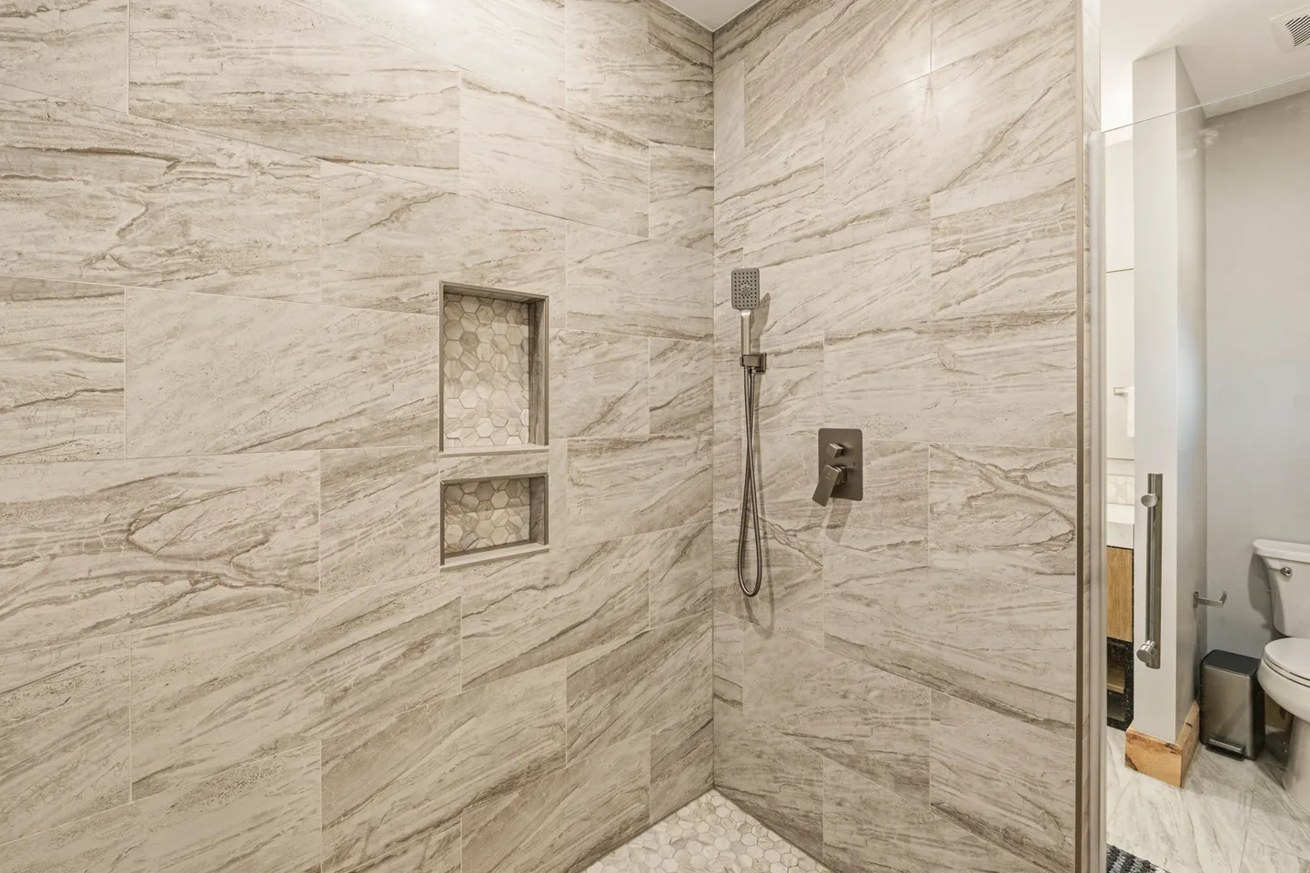 Walk-in shower with beige veined wall tiles, a mosaic tile niche, and dark fixtures next to a toilet.