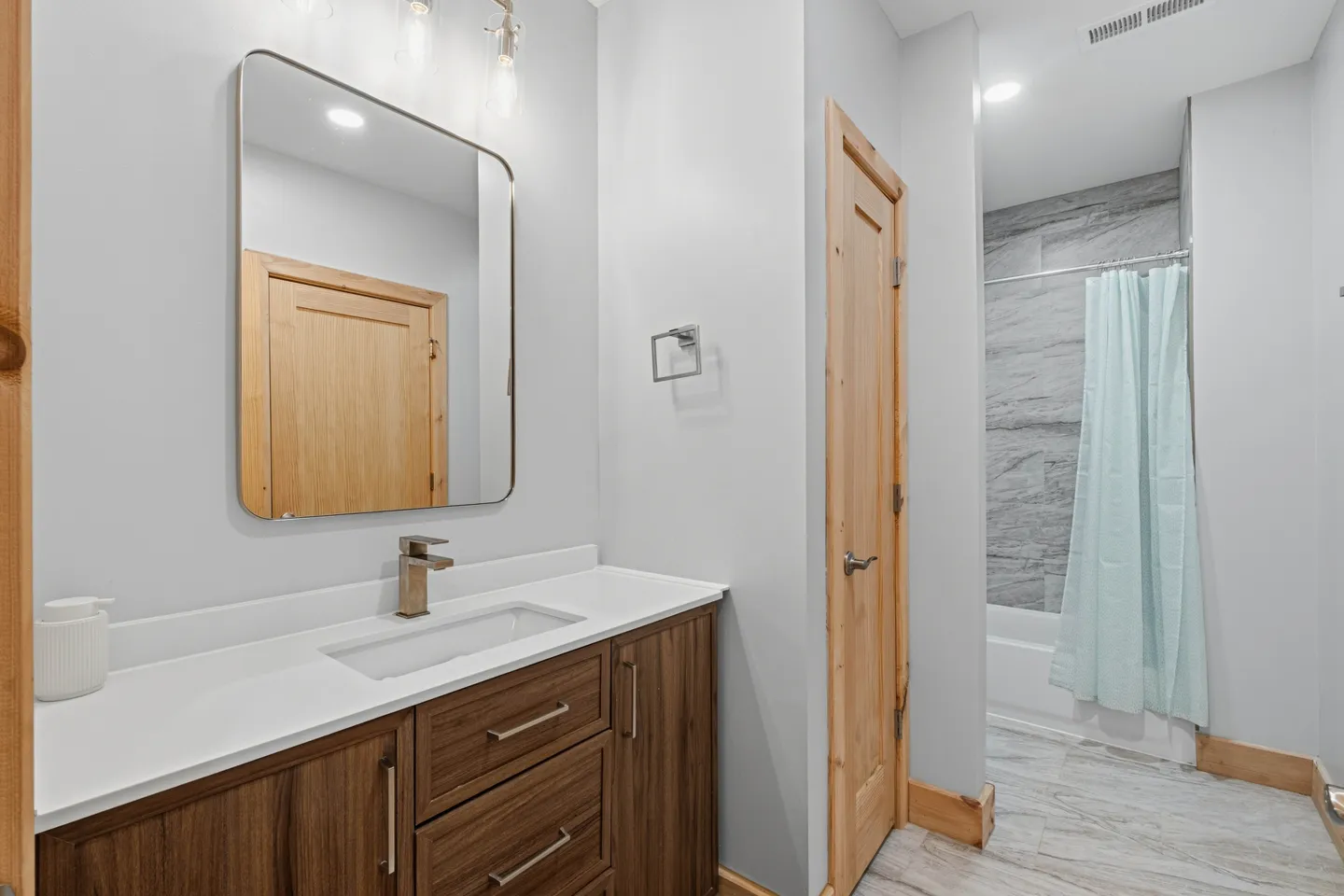 Bathroom with a wood vanity, white countertop, rectangular mirror, and adjacent bathtub-shower with a light blue curtain.