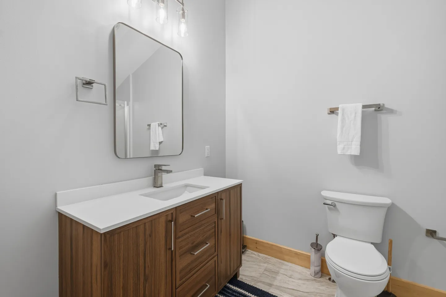 Bathroom with a wood-finish vanity, white countertop, modern mirror, and toilet beside a light gray wall.