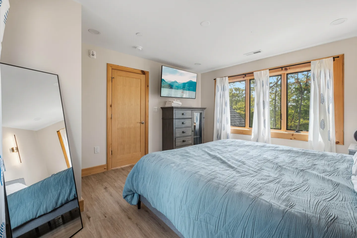 Bedroom with a bed made with blue bedding, a full-length mirror, a tall dresser, and windows looking out to trees.