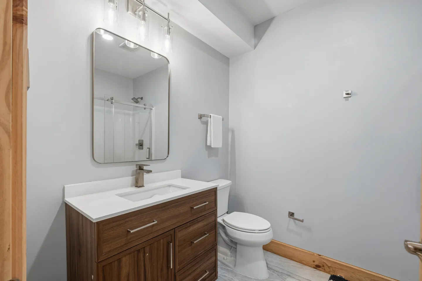 Bathroom featuring a dark wood vanity with a white countertop, modern mirror, toilet, and a glass-enclosed shower seen in the mirror reflection.
