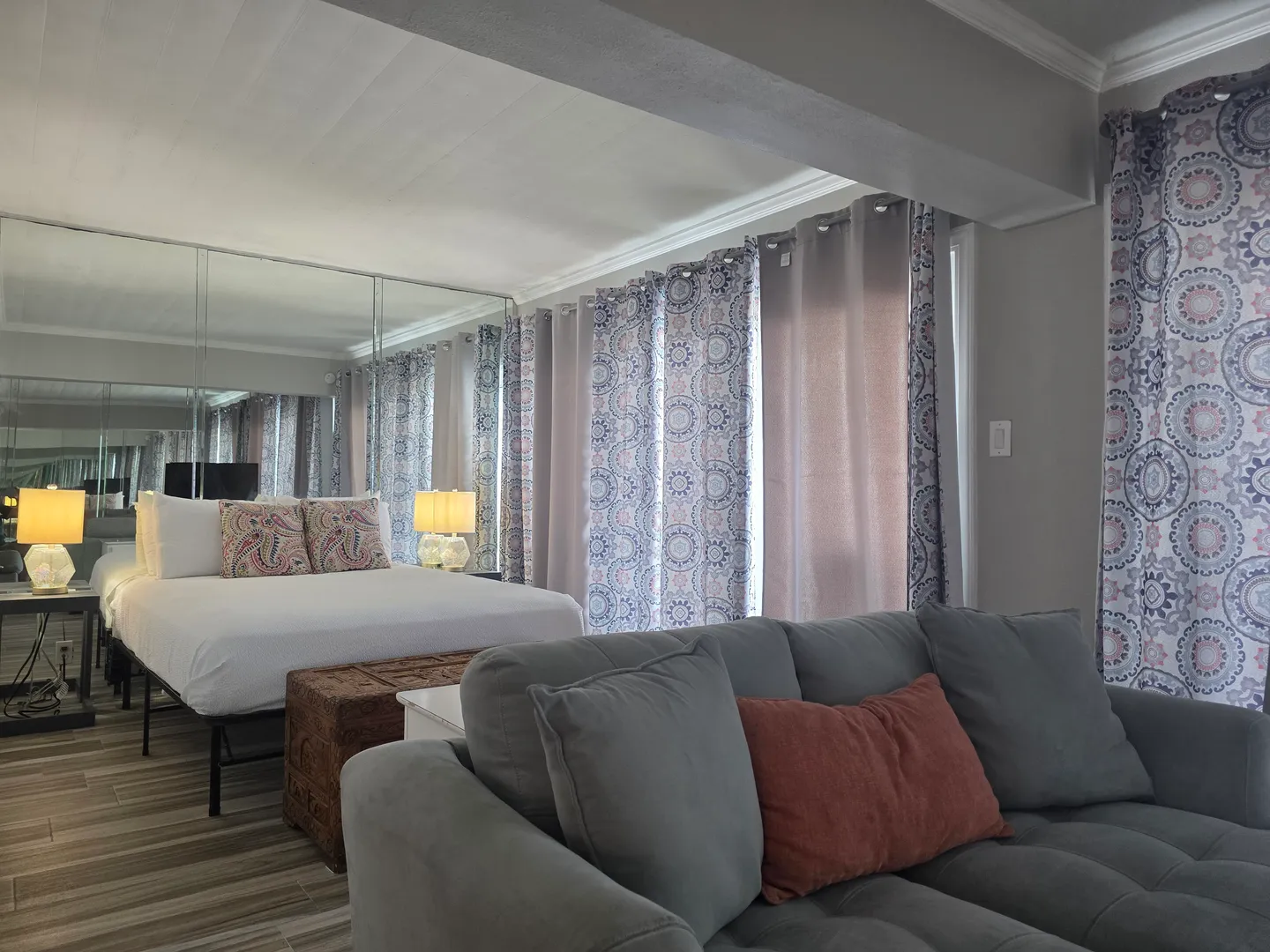 Bedroom and living area with a bed, mirrored wall, patterned curtains, and a gray sofa.