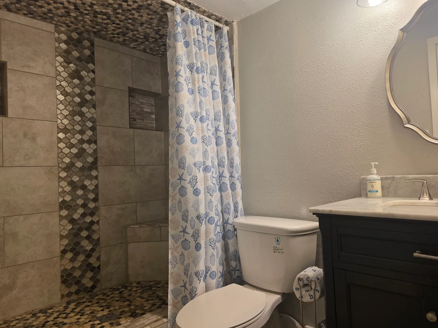 Bathroom showing a tiled walk-in shower with sea-themed curtain, a white toilet, and a dark vanity with a round mirror.