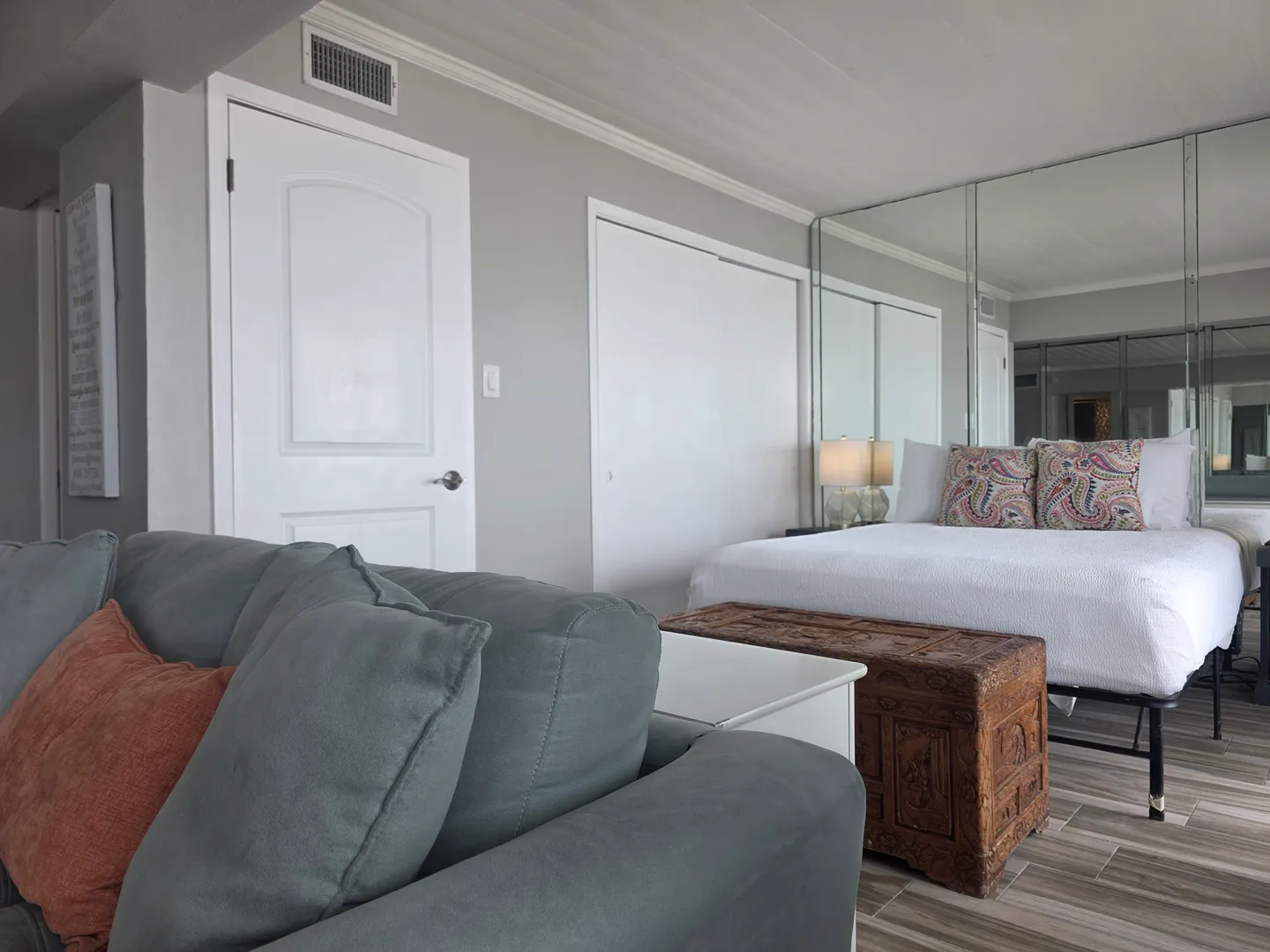 Bedroom area featuring a bed with patterned throw pillows, a large mirrored wall, a carved wooden chest, and an adjacent gray sofa.