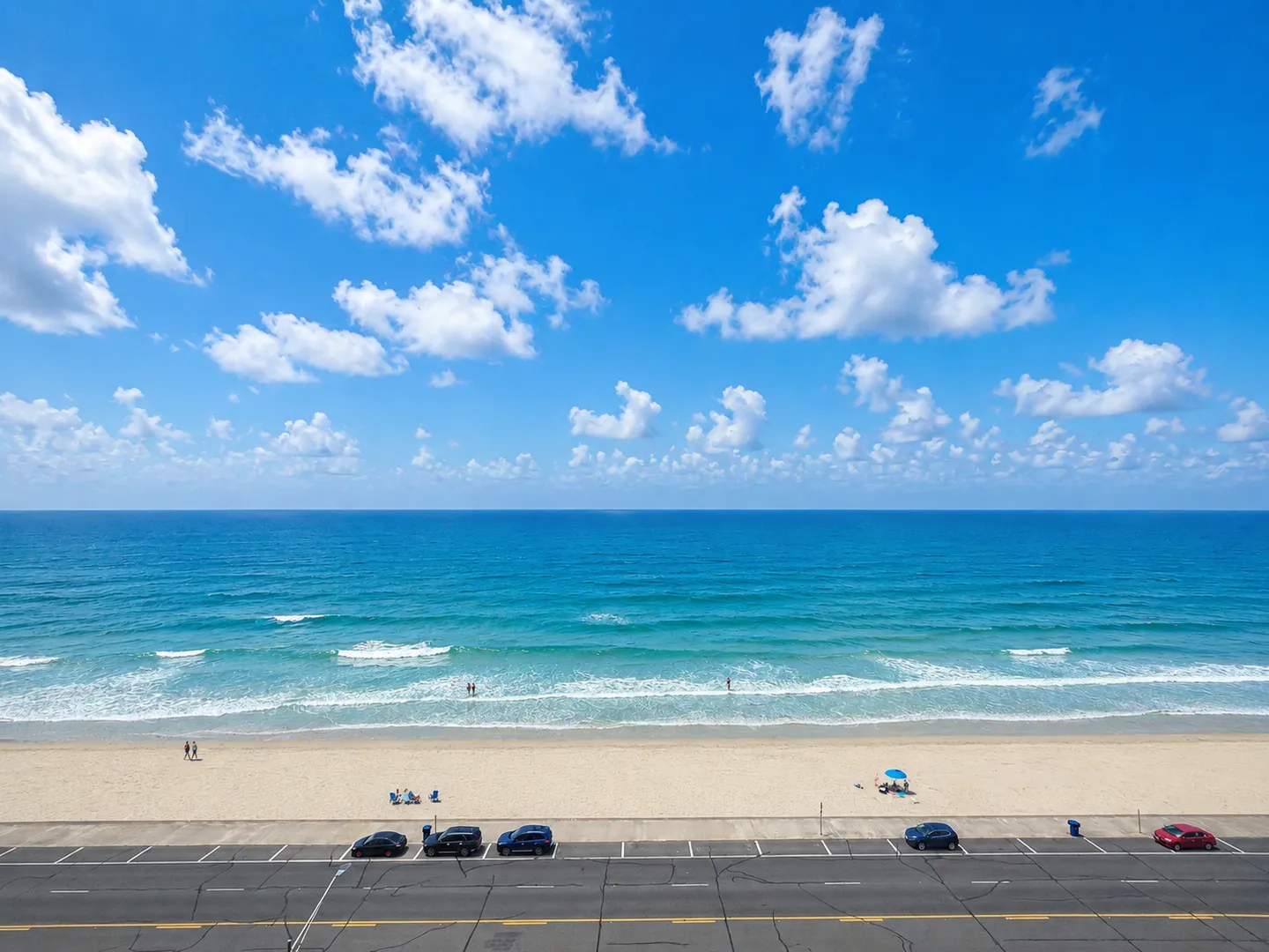 Elevated view overlooking a paved road, sandy beach, and turquoise ocean under a bright blue sky with scattered clouds.