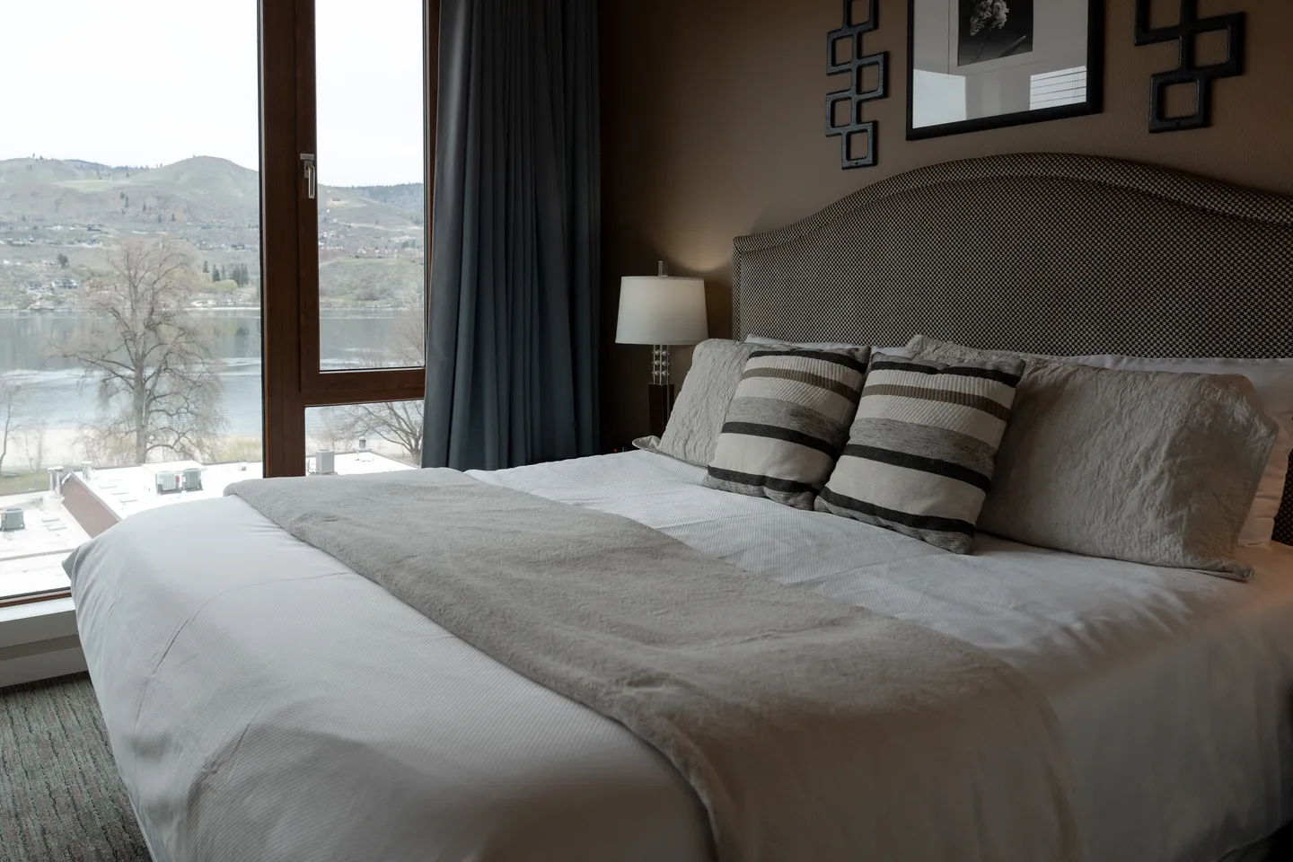 Bedroom featuring a made bed with an upholstered headboard next to a window overlooking a body of water and hills.