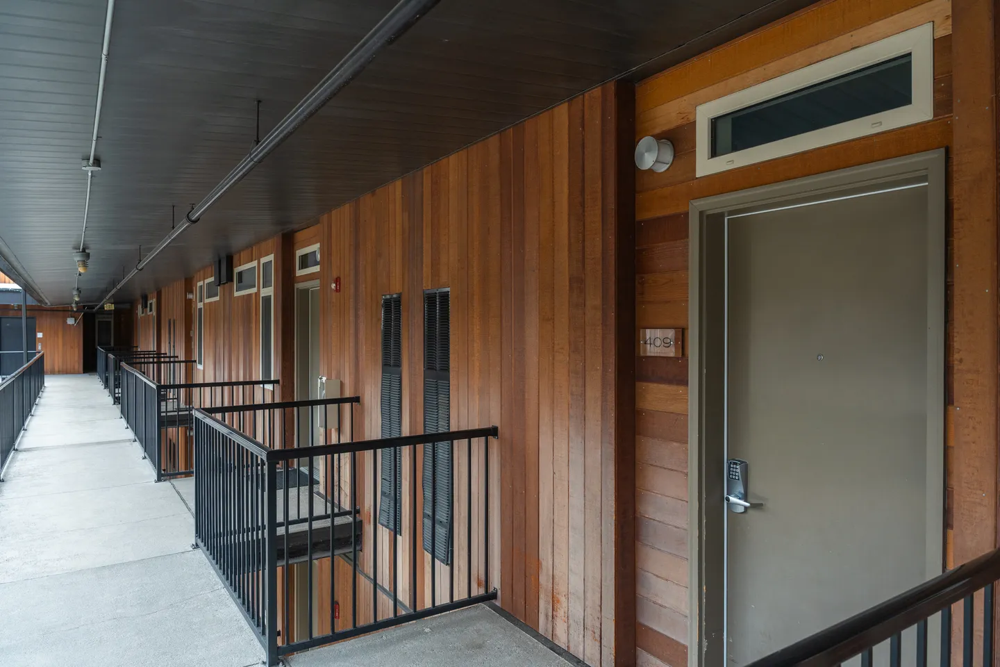 Exterior open-air walkway lined with vertical wood paneling, black metal railings, and unit entrance doors.