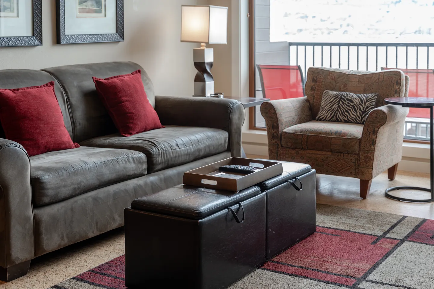 Living room featuring a dark sofa with red accent pillows, an upholstered armchair, storage ottomans as a coffee table, and a sliding glass door to a balcony.