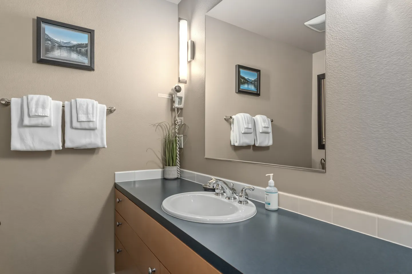 Bathroom vanity with a dark countertop, white drop-in sink, large frameless mirror, and folded white towels on a wall rack.