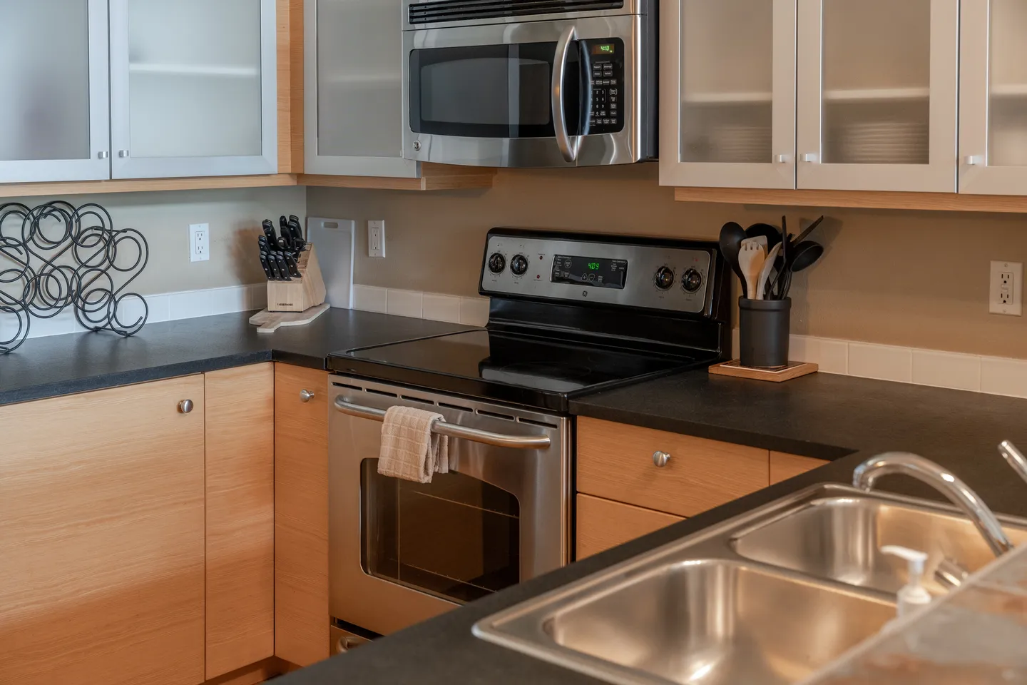 Close-up of a kitchen corner featuring an electric range, over-the-range microwave, dark countertops, and light wood cabinetry with frosted glass upper panels.