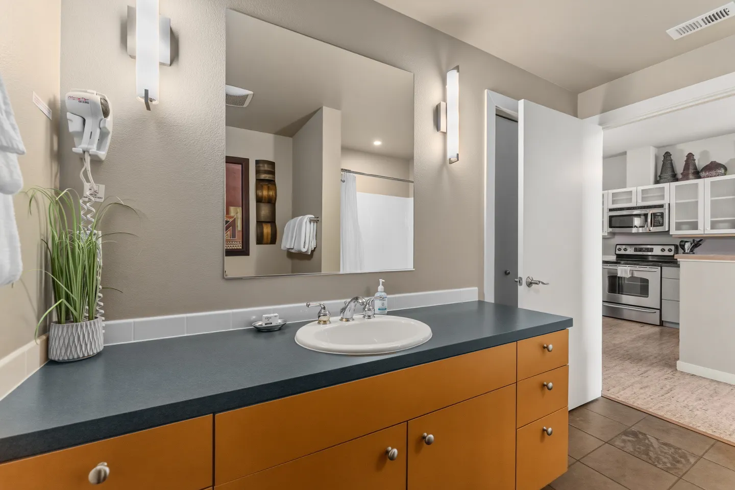 Bathroom vanity with a dark countertop, white sink, wall-mounted mirror, and an open doorway showing a view into the kitchen.