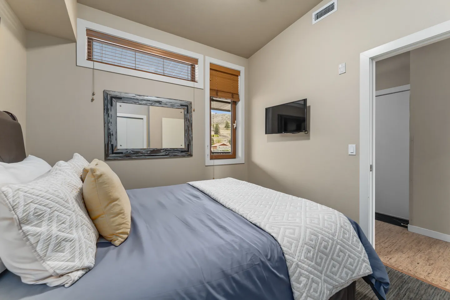 Bedroom with a made bed, wall-mounted television, rustic framed mirror, and windows with blinds.