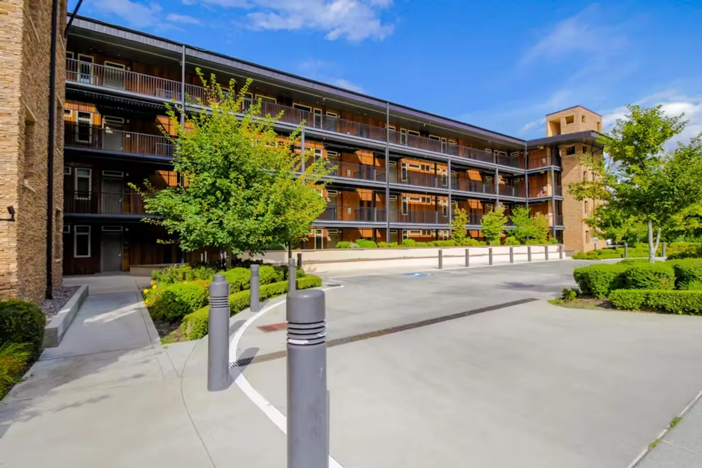 Exterior view of a multi-story modern apartment or condominium building with exterior walkways, surrounded by concrete paths, bollards, and manicured landscaping under a blue sky.