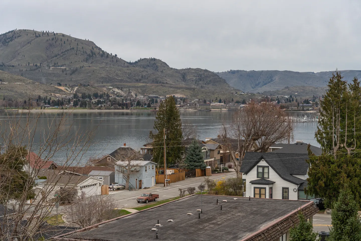 Elevated view overlooking houses, a street, and a calm lake bordered by rolling hills under an overcast sky.