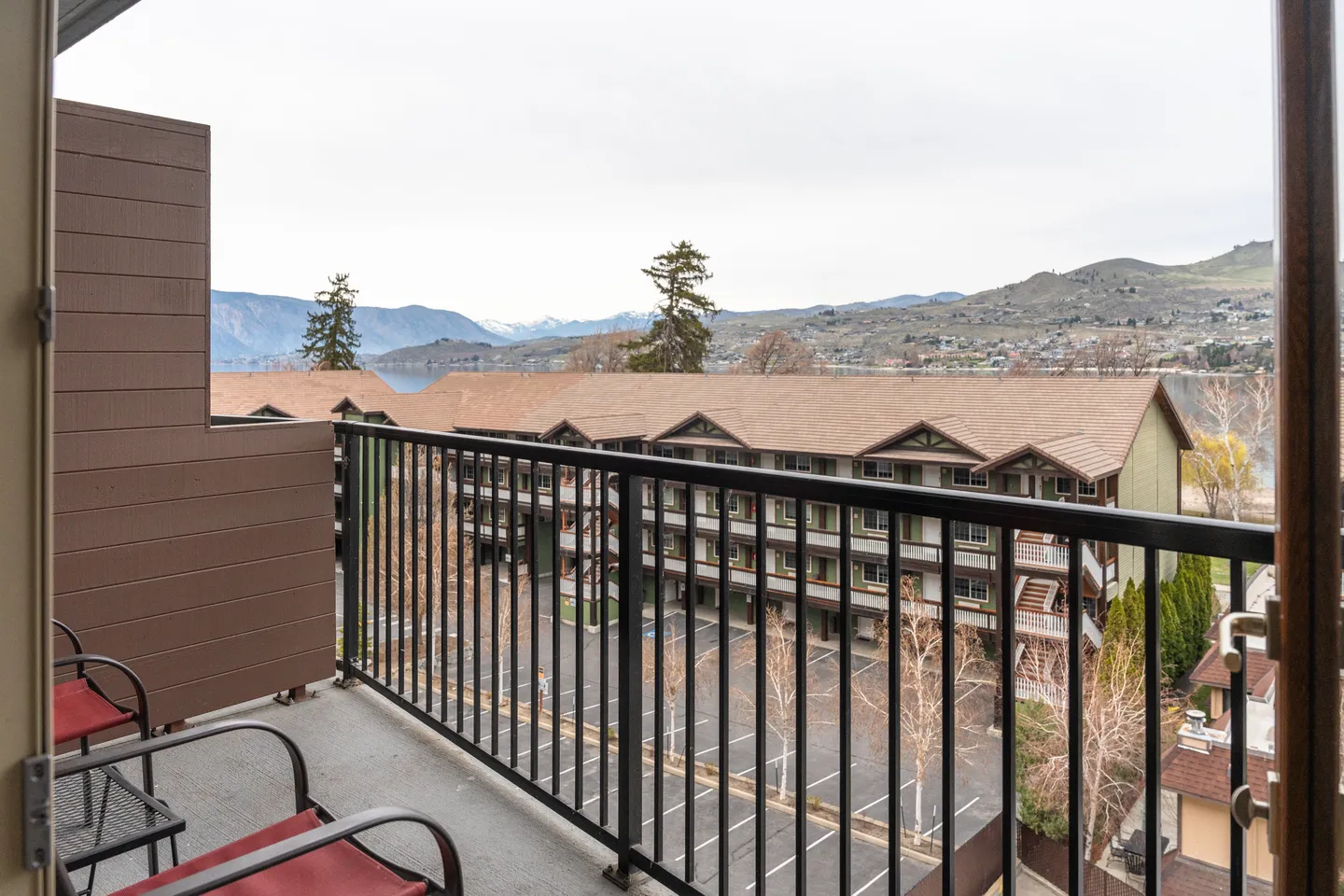 Balcony with black metal railings and patio chairs looking out over neighboring buildings toward hills and water.