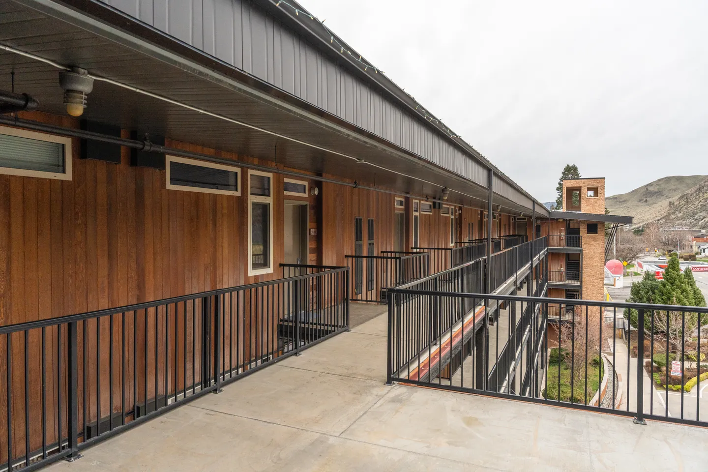 Exterior open-air walkway on an upper level of a multi-story building with wood-paneled walls and black metal railings.