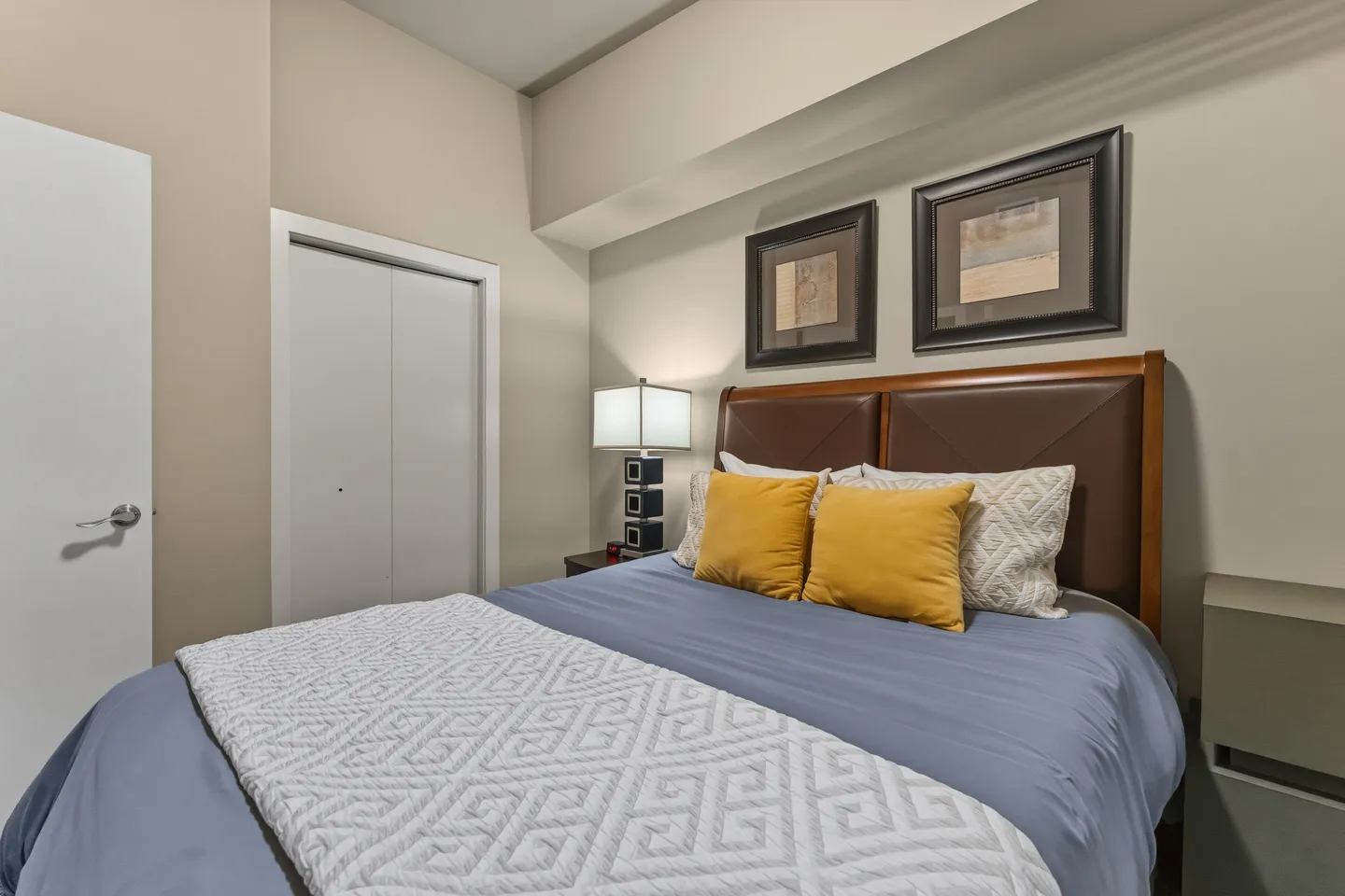 Bedroom featuring a made bed with brown headboard, yellow and patterned pillows, a bedside table with lamp, framed art, and a closet.