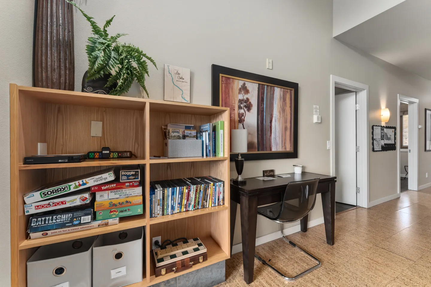 Hallway area with a wooden bookshelf stocked with board games, a small dark writing desk with a transparent chair, and doors leading to adjacent rooms.