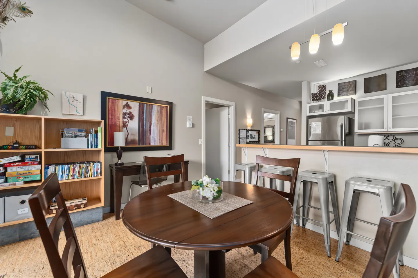 Dining area with a round wooden table, surrounded by wooden chairs, next to a bookcase and a kitchen breakfast bar with metal barstools.