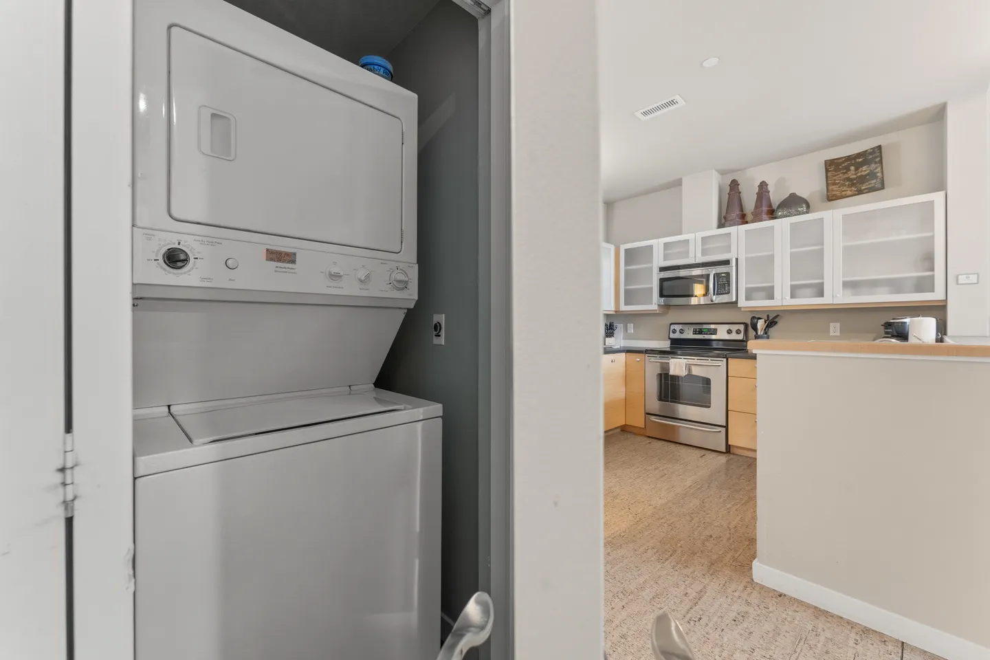 A stacked white washer and dryer unit inside a closet doorway, with the kitchen visible in the background.