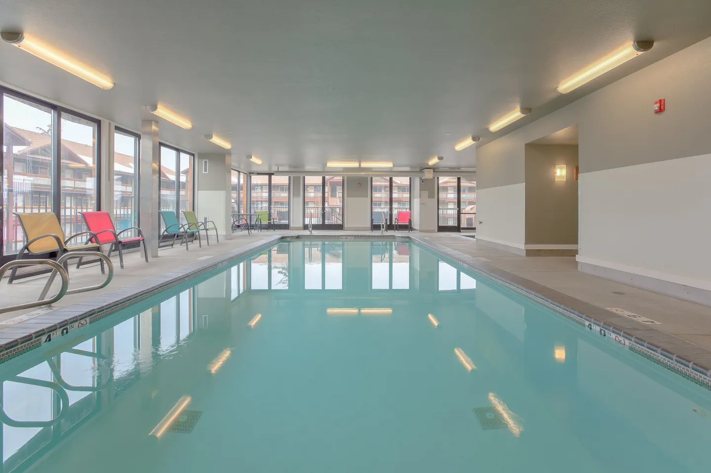 Indoor rectangular swimming pool lined with colorful lounge chairs and large windows looking outside.