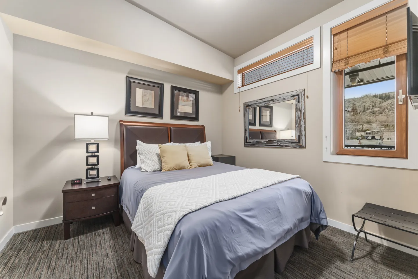 Bedroom with a made bed, headboard, nightstand with a lamp, wall mirror, and two windows.