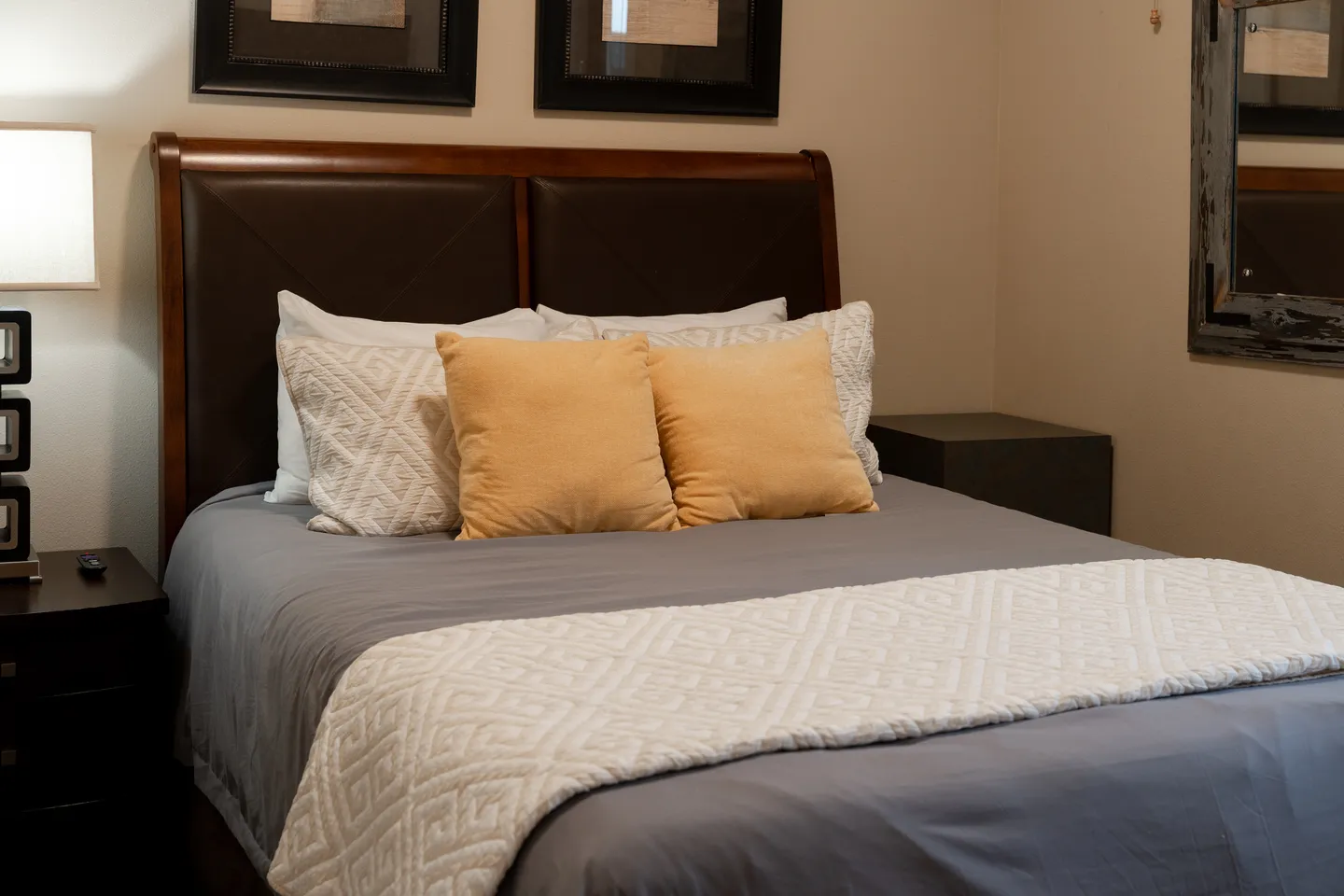 Bedroom featuring a made bed with a dark wood and leather headboard, yellow and patterned pillows, and a nightstand with a lamp.