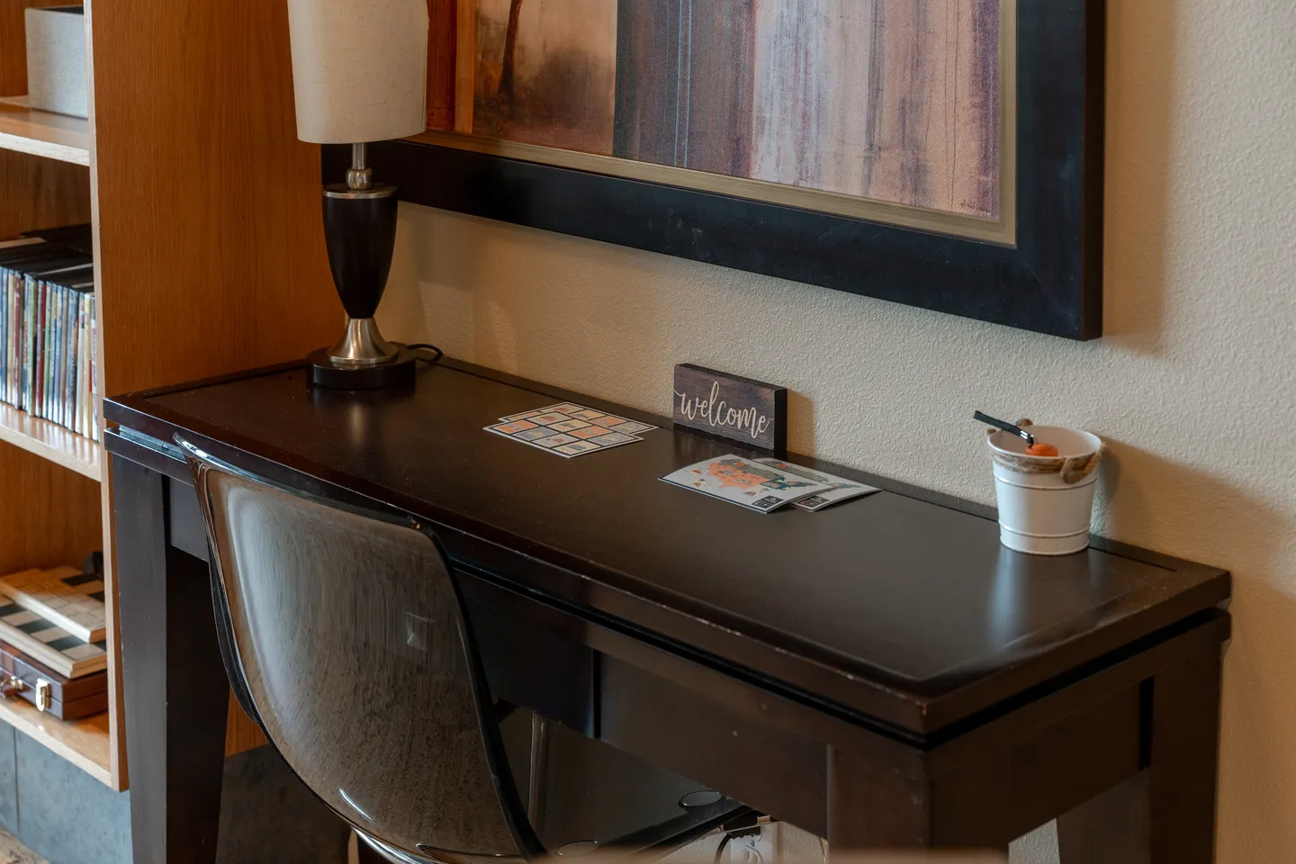 Dark wooden desk with a translucent chair, table lamp, small welcome sign, and framed artwork hanging above.