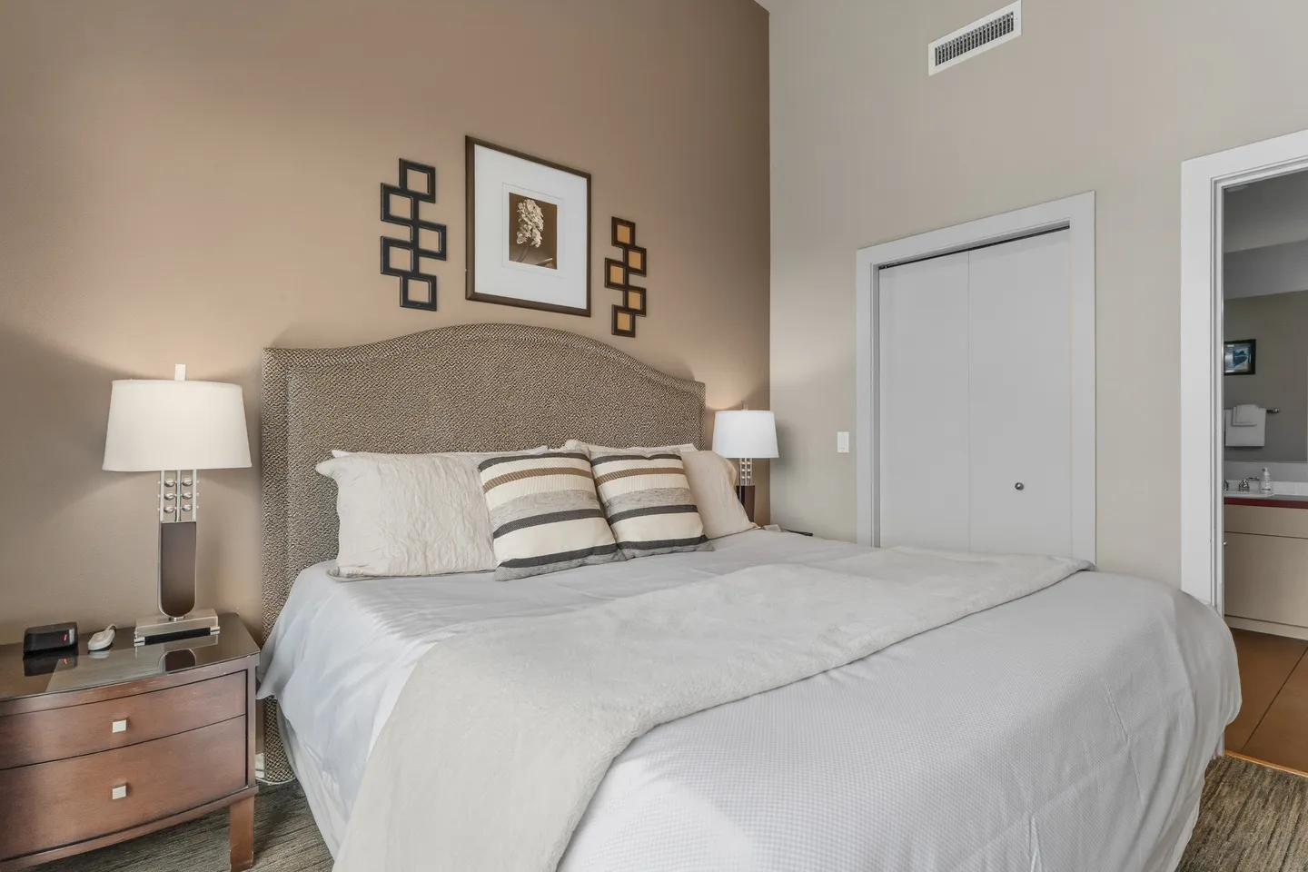 Bedroom with an upholstered headboard, dressed bed, side tables with lamps, closet, and view into an adjacent bathroom.