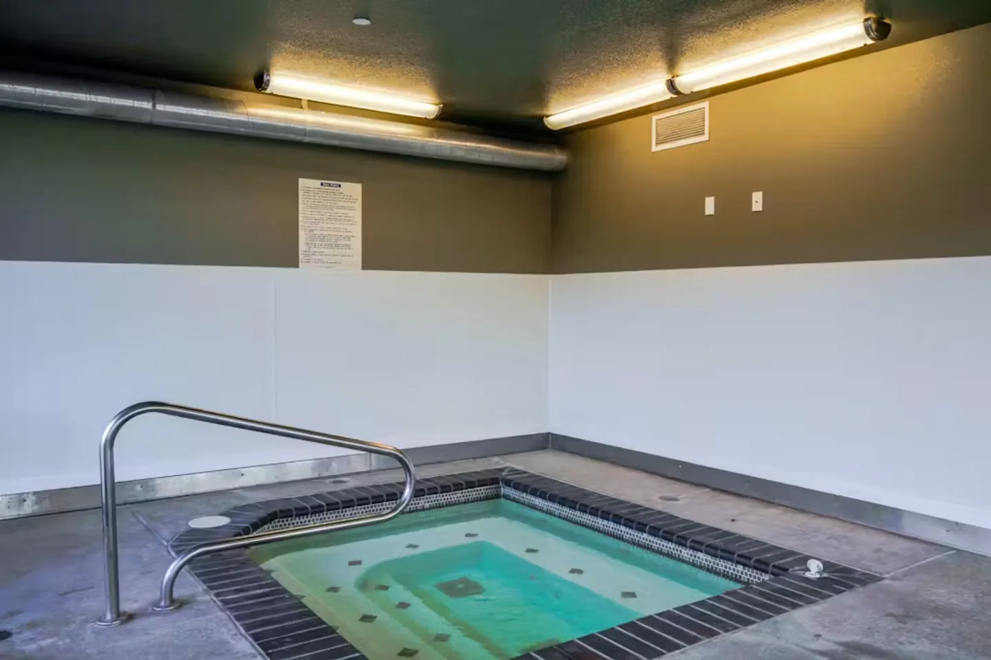 Indoor hot tub with metal handrail surrounded by tiled edge and concrete flooring under overhead fluorescent lights.