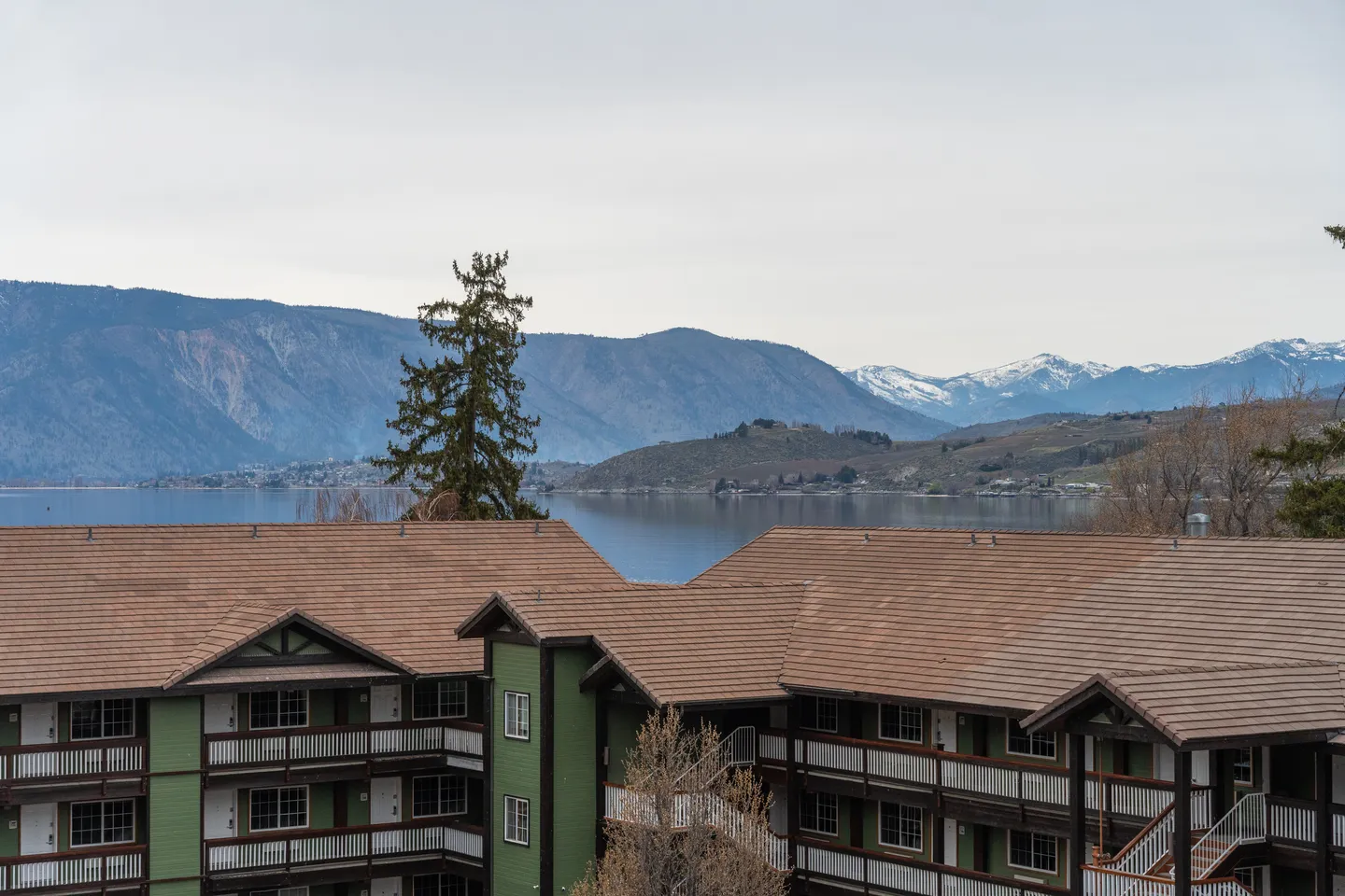 Exterior view of multi-story buildings with balconies, overlooking a lake and distant snow-capped mountains.