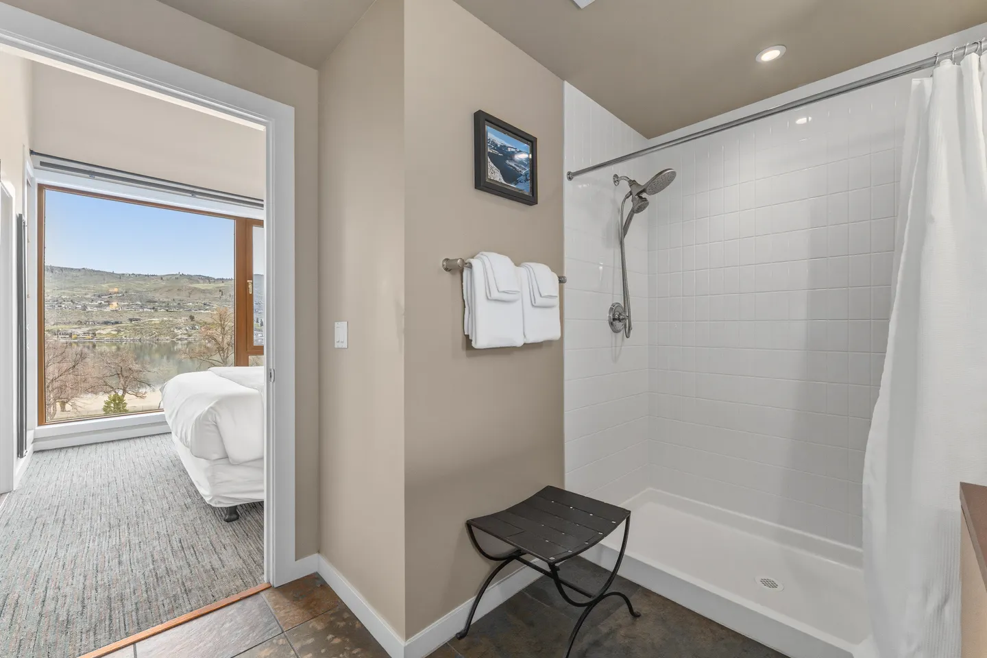 Bathroom with a white tiled walk-in shower and a doorway leading into a carpeted bedroom with large windows.