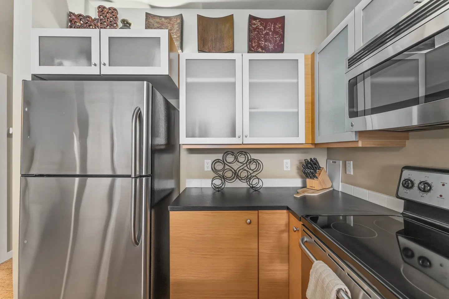 Kitchen corner featuring stainless steel refrigerator, black countertops, wood and frosted glass cabinets, and an electric range with microwave.