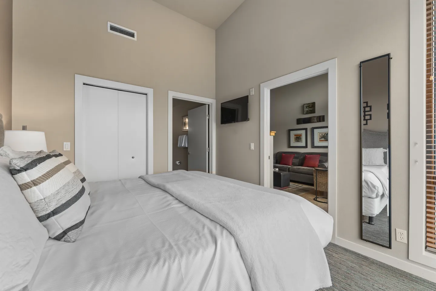 Bedroom with vaulted ceiling, bed dressed in white linens, wall-mounted television, and doorways leading to a bathroom and living area.