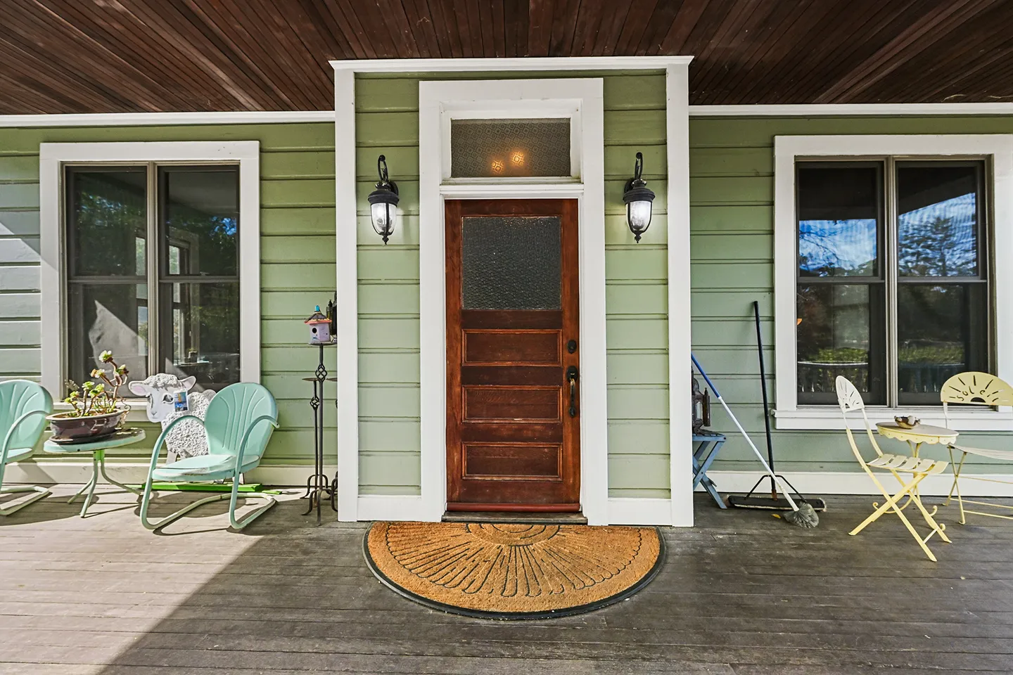 Front porch of a green house with a wooden entry door flanked by sconces, chairs, and potted plants.