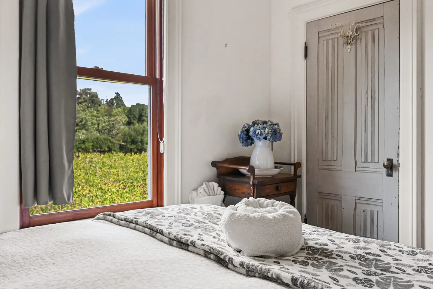 Bedroom detail with a bed in the foreground, rolled towel, wooden bedside table, and window overlooking a vineyard.