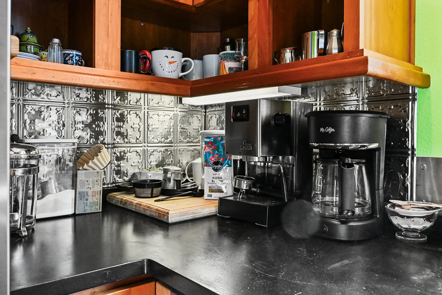 A kitchen countertop coffee station with an espresso machine, drip coffee maker, coffee supplies, and upper open shelves holding mugs.